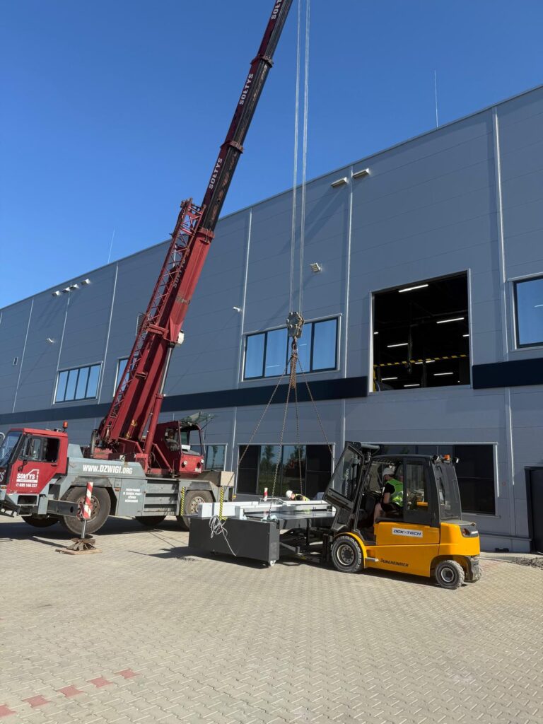 Dźwig i&nbsp;wózek widłowy DGK-Tech wprowadzają urządzenie przez otwór montażowy hali produkcyjnej